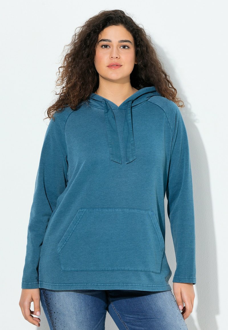 Donna con capelli ricci che indossa una felpa con cappuccio blu e jeans blu, in piedi contro uno sfondo chiaro e uniforme.
