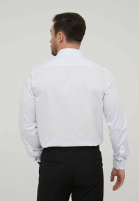 Chemise blanche à manches longues avec un col classique, texture lisse et poignets à boutons, associée à un pantalon noir, vue de dos.