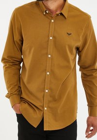 Koszula z guzikami w kolorze musztardowym, z kołnierzykiem, długimi rękawami oraz małym haftowanym logo na piersi. Wykonana z gładkiego materiału.