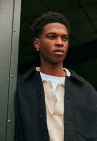 Black corduroy jacket over a beige knit sweater with a V-neck. The fabric appears textured, and the jacket features button closures and a pocket.