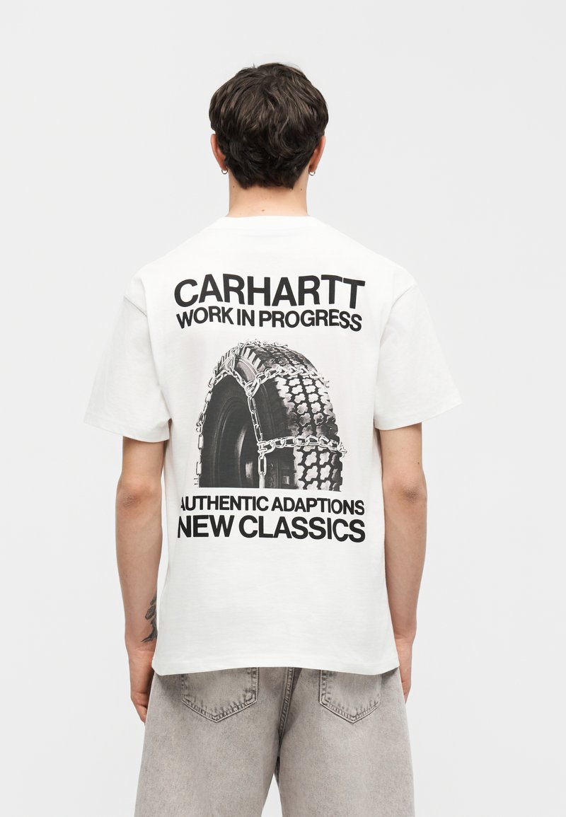 Persona che indossa una t-shirt bianca Carhartt con grafica nera di pneumatico e catena e testo in grassetto sulla schiena, abbinata a pantaloncini grigio chiaro.