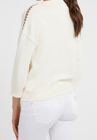 Donna che indossa un maglione lavorato a maglia di colore avorio con dettagli a catena sulla spalla e jeans bianchi aderenti, vista da dietro.