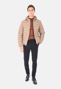 Uomo che indossa una giacca a imbottitura beige, una camicia marrone, pantaloni neri e stivali neri, in piedi contro uno sfondo bianco.