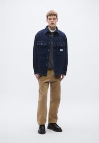 Μπλε σκούρο πουκάμισο-σακάκι από τζιν με δύο μπροστινές τσέπες, συνδυασμένο με καφέ παντελόνι και μαύρα παπούτσια. Το μοντέλο στέκεται μπροστά σε λευκό απλό φόντο.