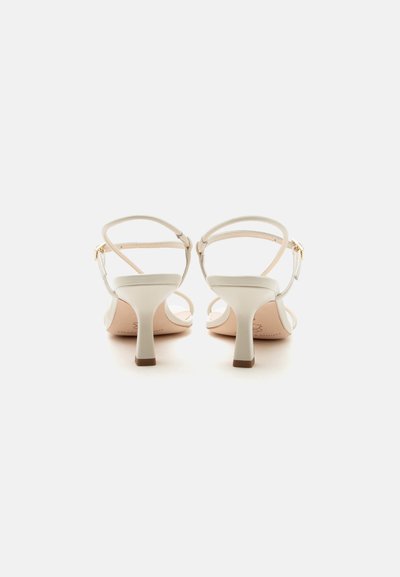 Een paar beige open-teen sandalen met hoge hakken, enkelriempjes en gouden gespen, gezien van achteren op een witte achtergrond.