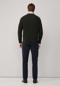 Maglione in maglia a treccia verde scuro con scollo rotondo, abbinato a pantaloni blu scuro e scarpe marroni, visto da dietro.