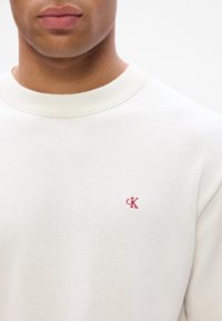 Švarkas apykakle off-white spalvos audiniu su tekstūruotu paviršiumi; turi mažą raudoną išsiuvinėtą "ck" logotipą kairėje krūtinės pusėje.