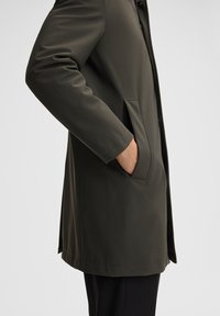 Cappotto impermeabile verde oliva con taglio dritto, tasche laterali e finitura liscia. Presenta una chiusura a bottone e un design con colletto alto.