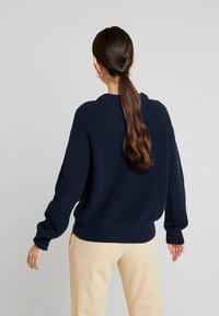 Maglione lavorato a maglia blu navy con una texture a costine, vestibilità ampia, maniche larghe e scollatura rotonda, abbinato a pantaloni beige chiaro.