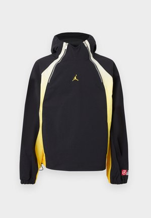 Svart og gul hettejakke med glidelåser foran nær kragen, elastiske mansjetter og en liten gul logo midt på brystet.