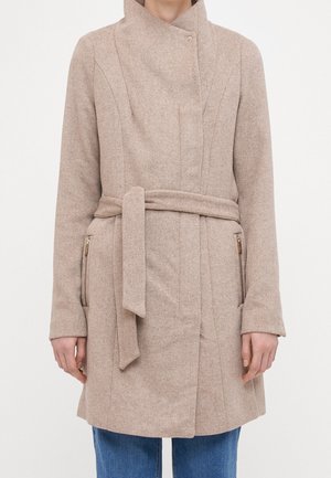 Osoba ubrana w beżowy wełniany płaszcz z paskiem i kieszeniami na zamek, do tego niebieskie dżinsy, na jednolitym białym tle.