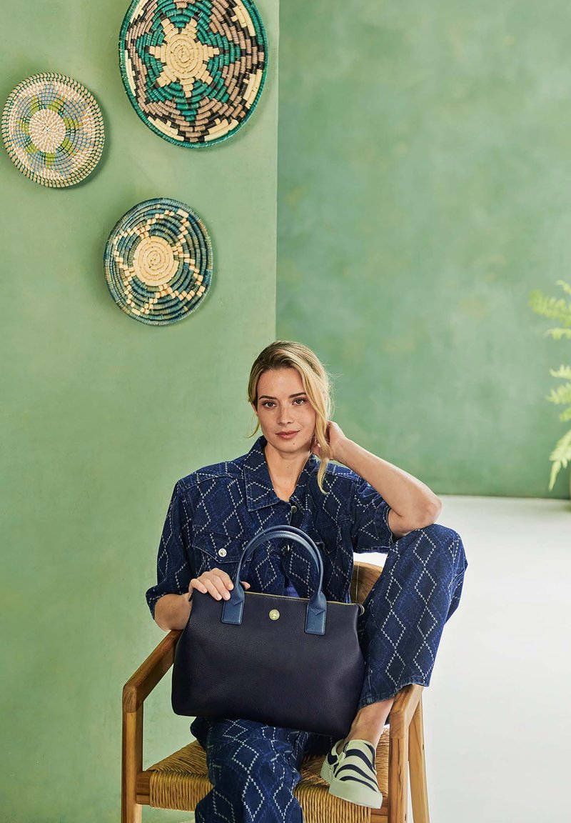 Marineblaue Lederhandtasche mit strukturiertem Design, doppelten Griffen und einem runden Emblem. Das Modell in passendem gemusterten Outfit sitzt auf einem Holzstuhl.