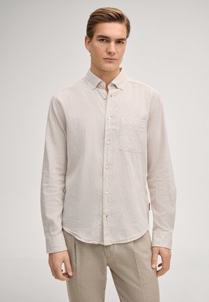 Junger Mann mit hellbraunen Haaren, der ein beiges langärmeliges Hemd mit Knöpfen und eine passende Hose vor einem schlichten Hintergrund trägt.