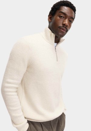 Jasno beżowy sweter zapinany na zamek z prążkowaną teksturą, wysokim kołnierzem i dopasowanymi rękawami, w połączeniu z ciemnymi spodniami. Miękka mieszanka wełny.