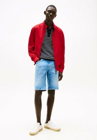 Giacca bomber leggera rossa sopra una polo a righe bianche e nere, abbinata a pantaloni corti di jeans azzurri e scarpe da ginnastica bianche.