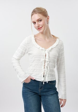 Femme aux cheveux blonds portant un haut blanc texturé à manches longues avec des nœuds noués à l'avant et un jean bleu, debout avec une main dans la poche.