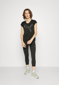 Černé sportovní tričko s bílým logem Puma, spárované s černými legínami po kotníky. Modelka má na sobě světle zelené sportovní boty a stojí na neutrálním pozadí.