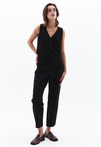 Zwarte vest met een diepe V-hals, knoopsluiting en zakken, gecombineerd met zwarte high-waisted broek en bruine loafers. Glad textuur.