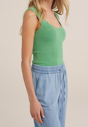 Vrouw draagt een nauwsluitend, mouwloos groen topje en lichtblauwe broek met trekkoord, staand tegen een neutrale achtergrond.