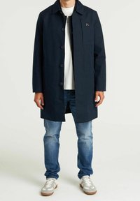 Hombre vestido con un abrigo azul marino abotonado, camiseta blanca, jeans azules y zapatillas blancas, de pie contra un fondo claro y liso.