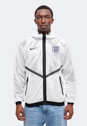 Mand iført en hvid net lynlåsjakke med Englands fodboldemblem og Nike-logo, kombineret med lyseblå jeans, vendt fremad.
