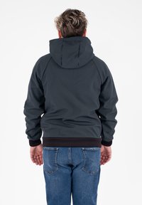 Persona con capelli ricci corti che indossa una giacca con cappuccio scura e jeans blu, in piedi di spalle su uno sfondo bianco.