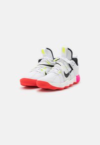 Zapatos deportivos blancos con una parte superior texturizada, acentos negros, detalles en amarillo neón y una suela en rosa y rojo. Presentan cordones y correas ajustables.