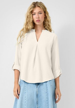 Jeune femme aux longs cheveux blonds bouclés portant un chemisier crème aux manches retroussées et une jupe en denim bleu clair sur un fond uni.