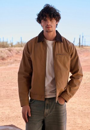 Junger Mann mit lockigem Haar trägt eine braune Jacke, weißes T-Shirt und Jeans und steht im Freien in einer trockenen, ländlichen Gegend mit Zäunen im Hintergrund.