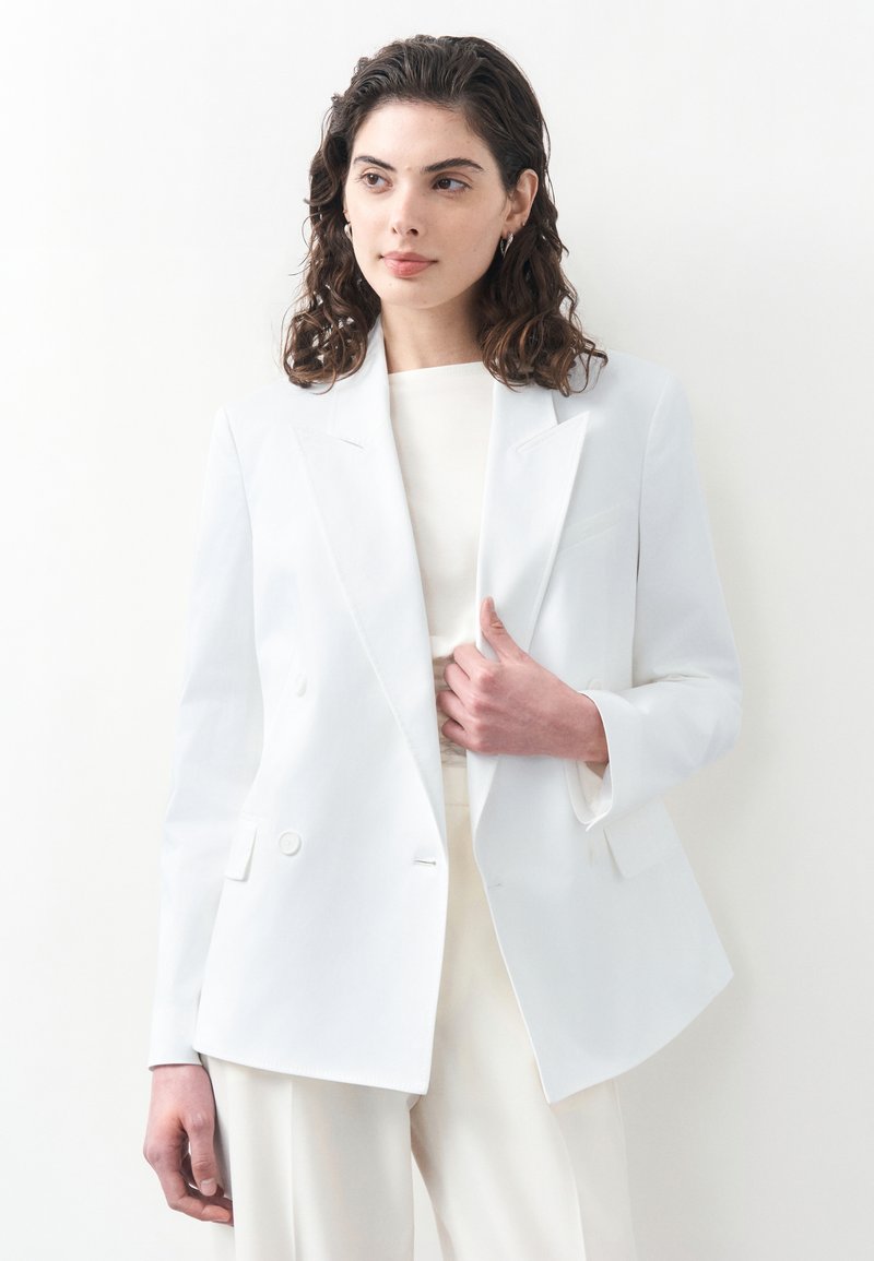 Femme aux cheveux bouclés foncés portant un blazer blanc par-dessus un haut et un pantalon blancs, tenant le revers du blazer, debout devant un fond blanc.