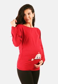 Rotes langärmeliges Stillshirt mit seitlicher Raffung, das eine Cartoon-Baby-Grafik zeigt, die von unten hervorblinzelt. Hergestellt aus weichem Stoff.