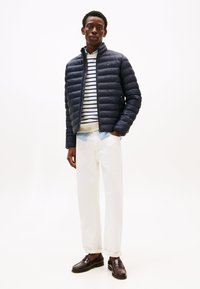 Doudoune bleu marine à motif matelassé, associée à un pull rayé blanc et bleu marine en dessous, pantalon blanc et mocassins foncés.