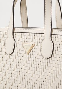 Geweven crèmekleurige handtas met zwarte accenten, gestructureerd ontwerp, korte handvatten en een gouden driehoekige logoplaat aan de voorkant.