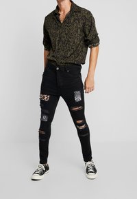 Jeans negros con múltiples parches y detalles desgastados, combinados con una camisa de un patrón verde oscuro. Presenta un ajuste casual y zapatillas negras.