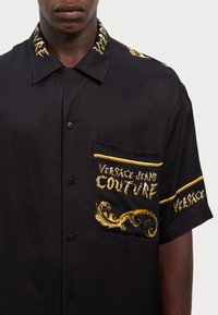 Black short-sleeved shirt featuring gold embroidery, with branding on the collar and chest pocket. Smooth fabric with a relaxed fit.