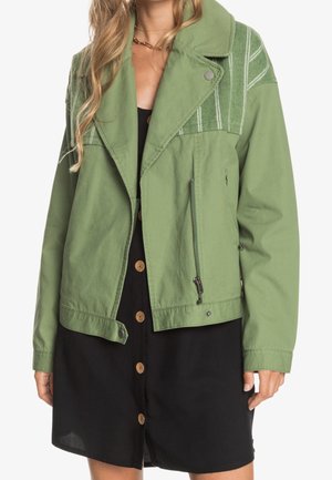 Kobieta w zielonej kurtce z paskowanymi detalami na ramionach, ubrana w czarną sukienkę zapinaną na guziki, stojąca na tle białego tła.