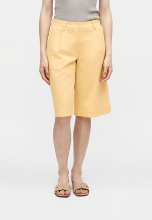 Persona che indossa pantaloncini gialli a vita alta, lunghezza al ginocchio con pieghe frontali, abbinati a sandali beige e top grigio senza maniche.
