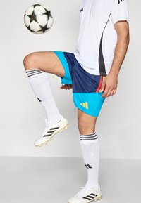 Conjunto de futebol com uma camisa branca, calções azuis e turquesa com detalhes vermelhos, e meias brancas com riscas pretas, combinados com chuteiras pretas e brancas.