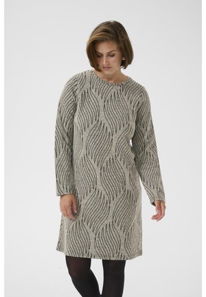 Vrouw met een grijze jurk met lange mouwen en zwarte golvende lijnpatronen, gecombineerd met zwarte panty's, staand tegen een effen achtergrond.