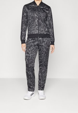 Personne portant une veste zippée à motif abstrait noir et gris et un pantalon assorti avec des baskets blanches Adidas, debout sur un sol blanc uni.