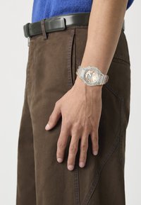 Montre transparente avec un cadran argenté et un bracelet transparent, portée sur une main reposant sur un pantalon marron avec des détails de surpiqûre contrastants.