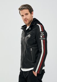 Zwarte leren motorjack met een capuchon, voorzien van patches en strepen in rood en wit langs de mouwen, met ritssluitingen aan de voorkant.