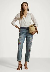 Sieviete, tērpta baltā pogas kreklā, ar krāsām noplūdušām džinsu biksēm, brūnām augstpapēžu kurpēm, tur rokās aušanas soma ar ādas siksnām.