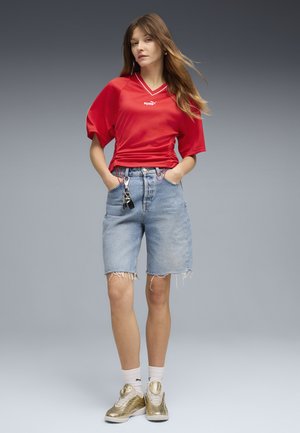 Mujer de pie con una camiseta roja de Puma, shorts vaqueros hasta la rodilla con llaves colgadas, calcetines blancos y zapatillas doradas sobre un fondo gris.