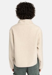 Una persona con cabello rizado oscuro viste una chaqueta de forro polar color crema y pantalones verdes, mostrada de espaldas contra un fondo claro y liso.