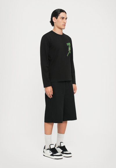 Svart langermet skjorte med grønt grafisk design, kombinert med svarte shorts til knelengde og svart-hvite joggesko. Grå sokker. Enkel tekstur.