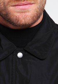 Černá bunda s úzkým límcem, stříbrným cvočkem a žebrovaným černým rolákem pod ní. Hladká, lesklá textura.