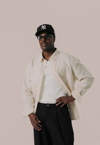 Stribet beige skjorte med knapper over en hvid tætsiddende top, parret med sorte bukser. Har en sort cap og briller, samt synlige detaljerne på lommen.