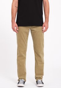 Pantalones de pana beige con piernas rectas, que presentan una textura de ribete vertical. Llevados con una camiseta negra y zapatillas negras.
