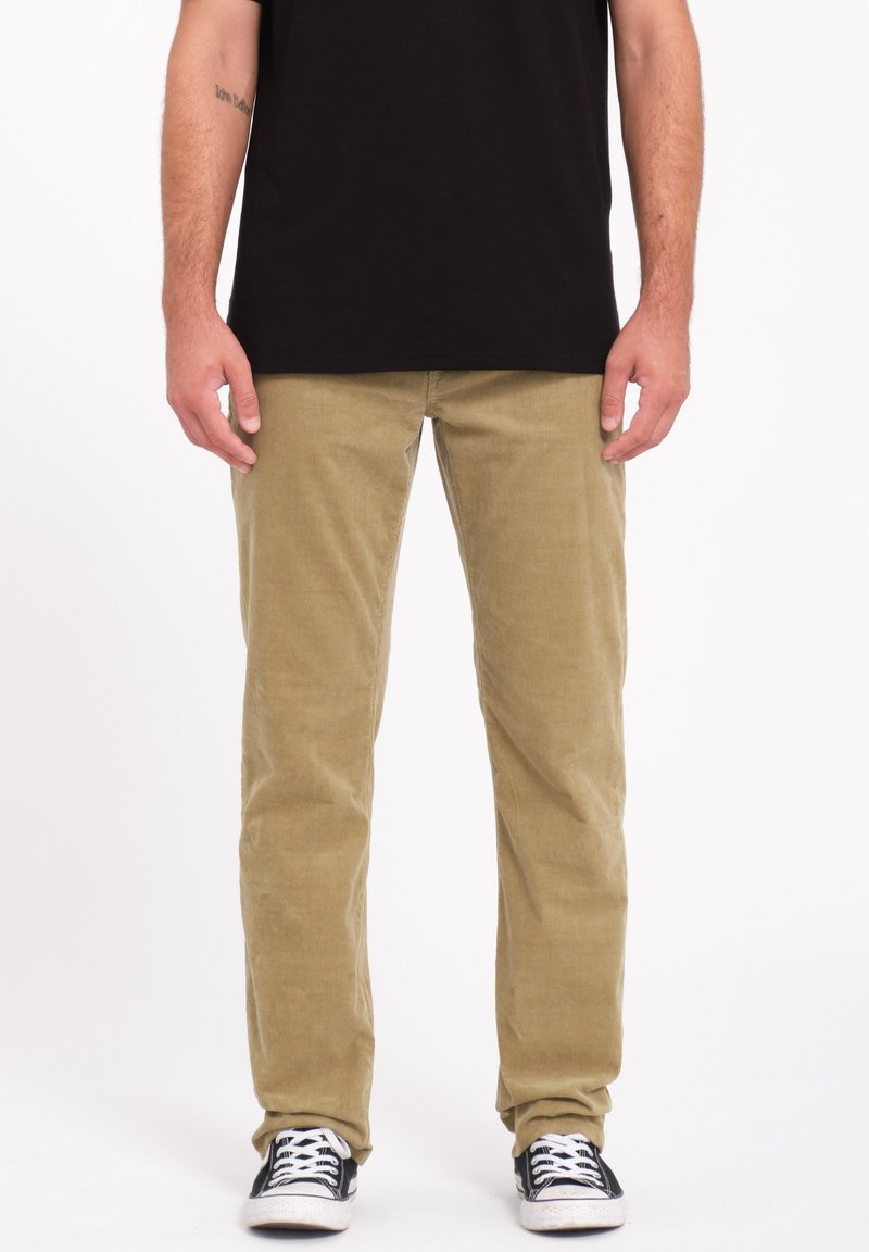 Pantalones de pana beige con piernas rectas, que presentan una textura de ribete vertical. Llevados con una camiseta negra y zapatillas negras.
